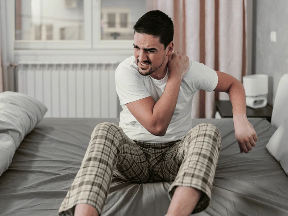 Man sitting on a bed and rubbing his shoulder.